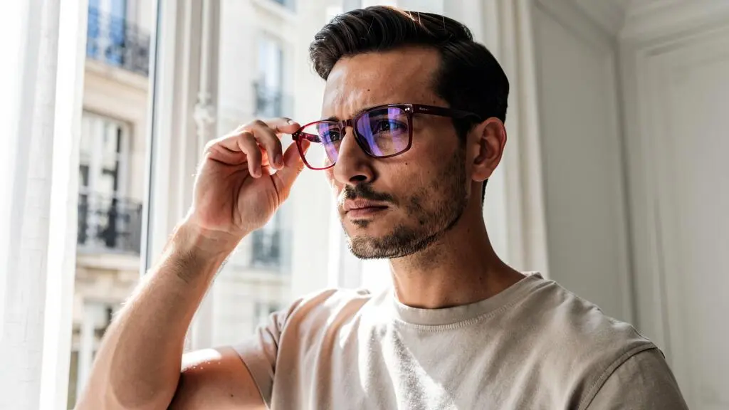 Un homme portant des lunettes de vue à monture bordeaux dans un appartement lumineux.